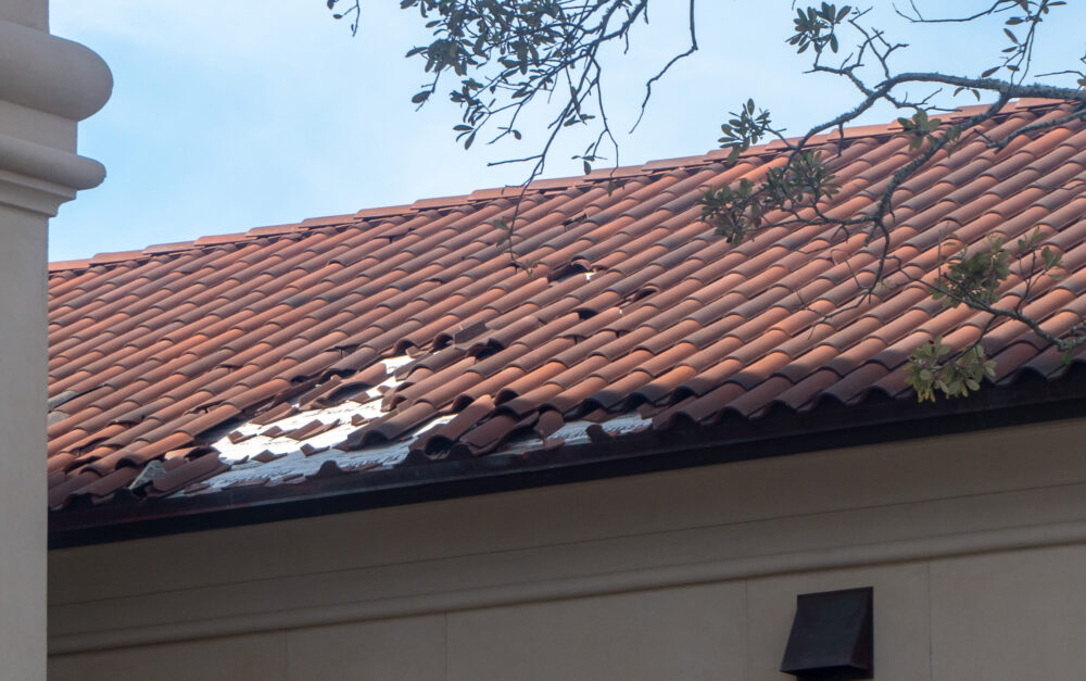 Red clay tile roof with missing tiles and exposed underlayment showing damage near the eave.