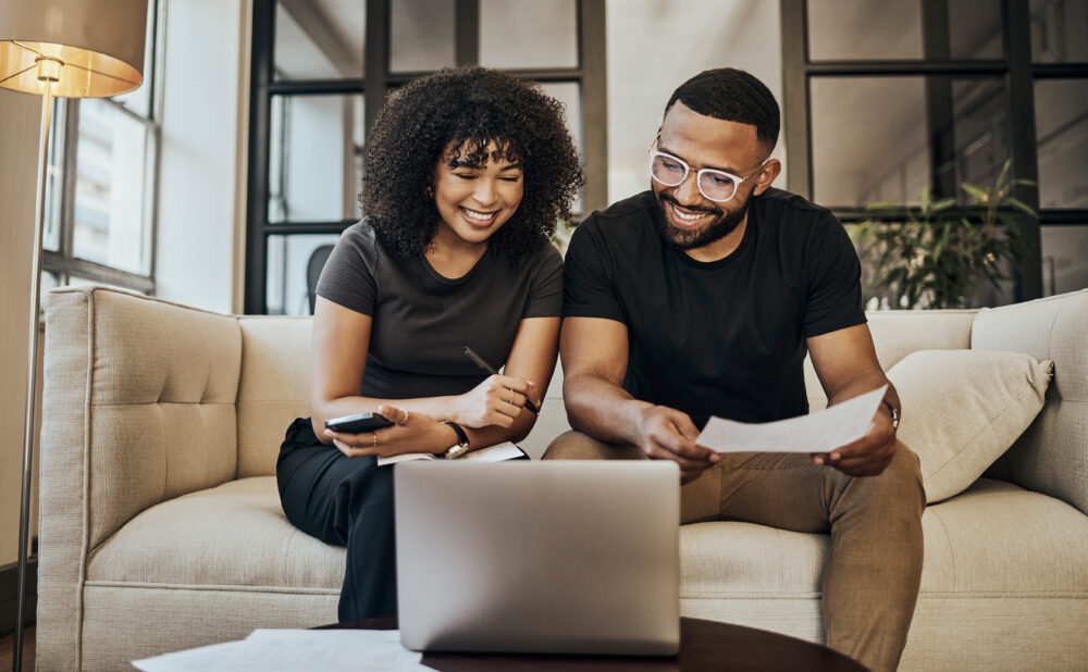 Couple sitting on a sofa, smiling while reviewing paperwork on a laptop in a modern living room.
