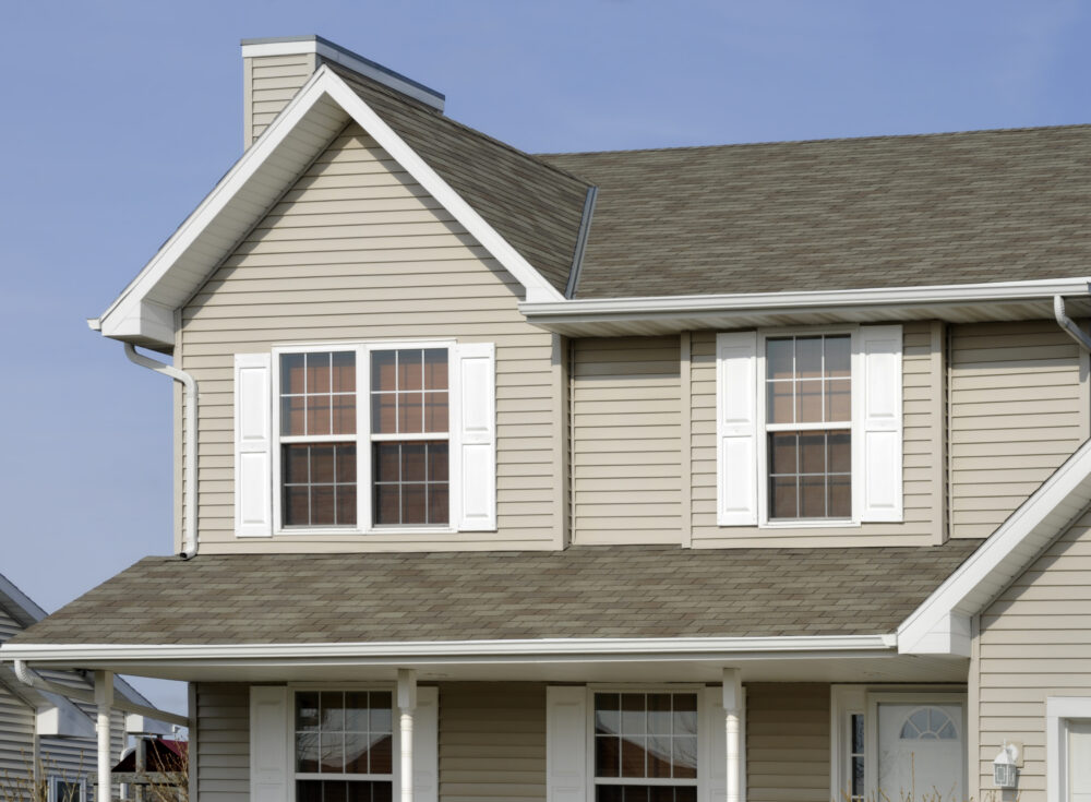 Two-story beige house with white shutters and a gray asphalt shingle roof under a clear blue sky.